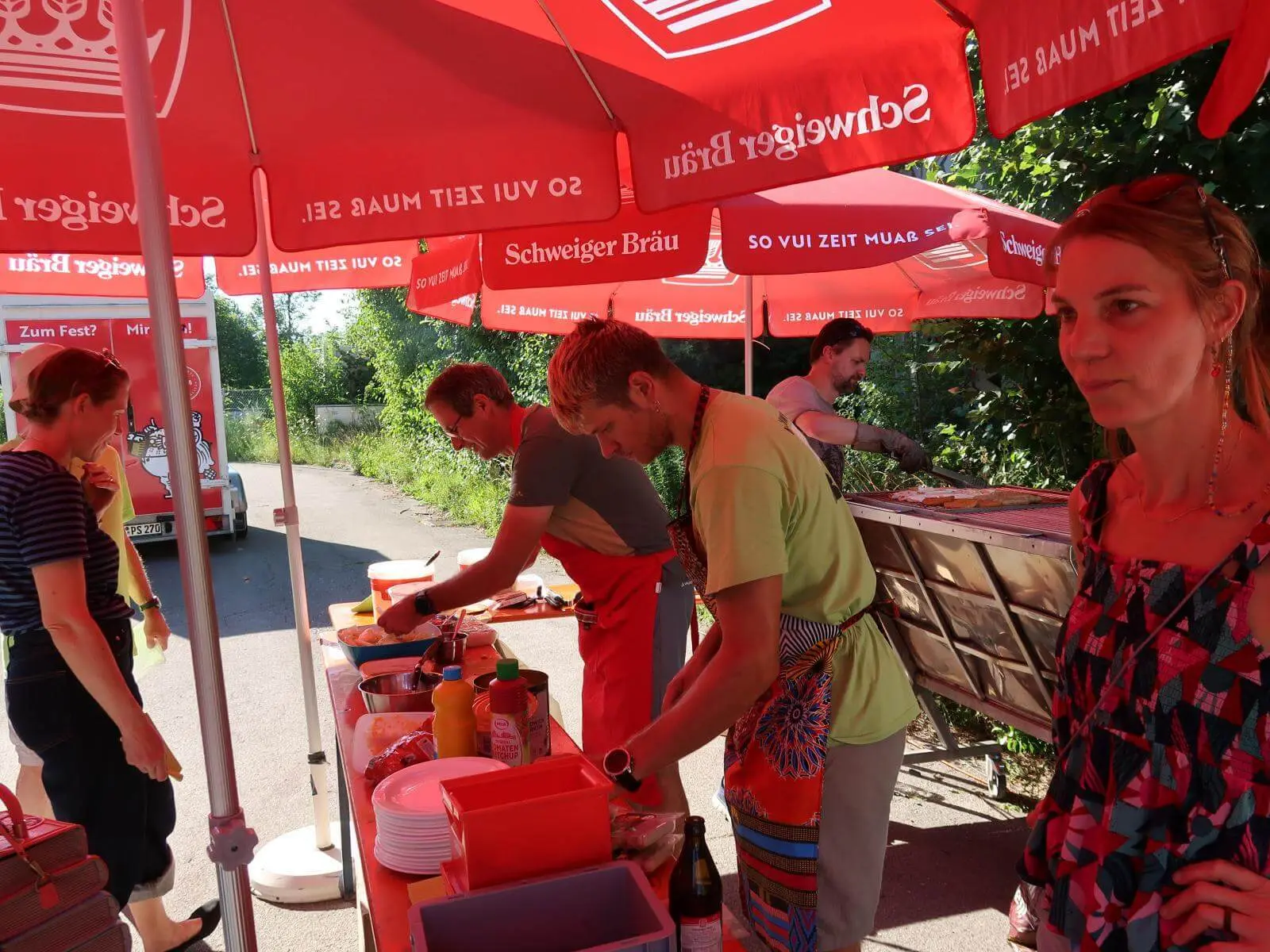 Team bereitet Essen am Grillstand unter roten Sonnenschirmen beim Sommerfest zu. | © DAV Markt Schwaben/Susanne Mayerl