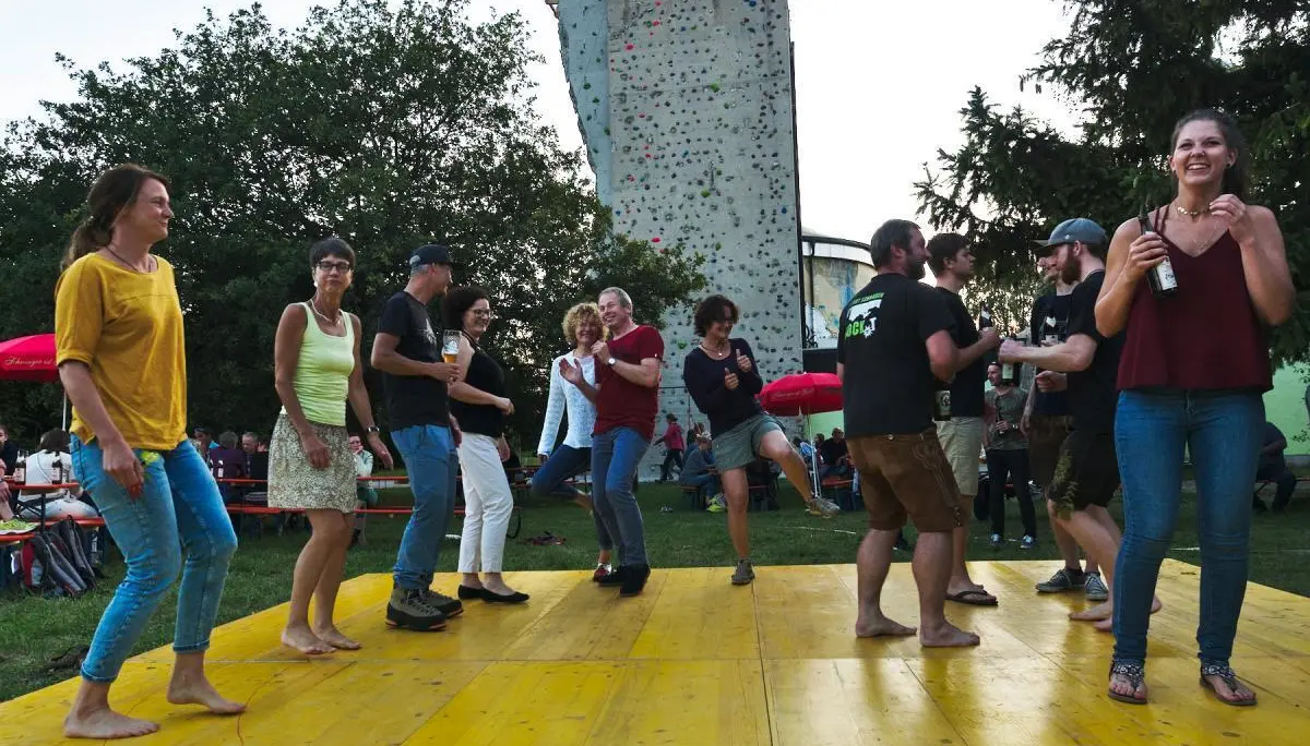 Viele Besucher sitzen an langen Tischen unter roten Sonnenschirmen, während im Hintergrund eine Bühne mit Livemusik zu sehen ist. | © DAV Markt Schwaben/Thomas Schäffler 2018