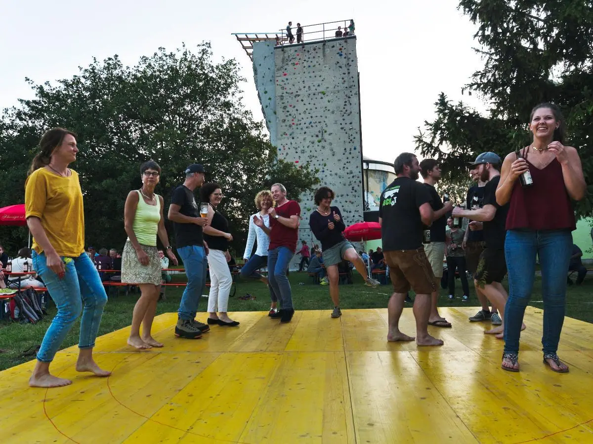 Viele Besucher sitzen an langen Tischen unter roten Sonnenschirmen, während im Hintergrund eine Bühne mit Livemusik zu sehen ist. | © DAV Markt Schwaben/Thomas Schäffler 2018