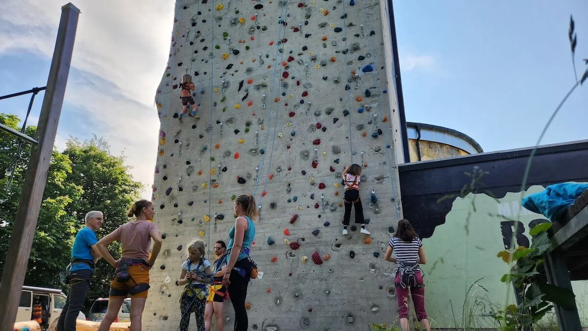 Familienklettern | © DAV Markt Schwaben Mehrere Personen klettern eine Outdoor-Kletterwand mit bunten Griffen hinauf. Zwei Kinder sind aktiv am Klettern, während andere daneben stehen, beobachten oder warten. | © DAV Markt Schwaben