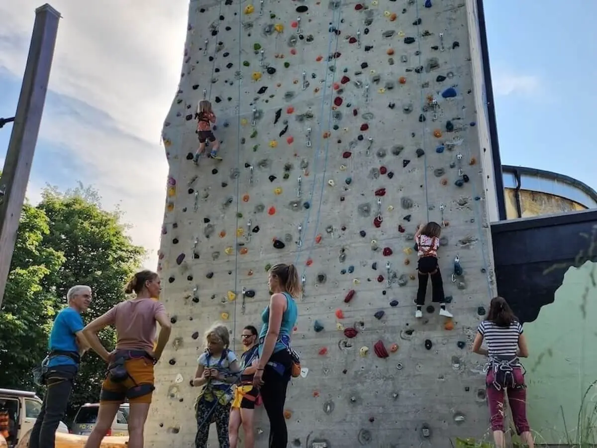 Mehrere Personen klettern eine Outdoor-Kletterwand mit bunten Griffen hinauf. Zwei Kinder sind aktiv am Klettern, während andere daneben stehen, beobachten oder warten. | © DAV Markt Schwaben