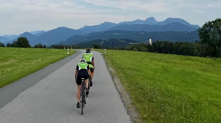 Zwei Radfahrer in grünen Trikots fahren hintereinander auf einer kurvigen Landstraße durch eine grüne Hügellandschaft, im Hintergrund Berge und ein Kirchturm unter leicht bewölktem Himmel. | © DAV Markt Schwaben