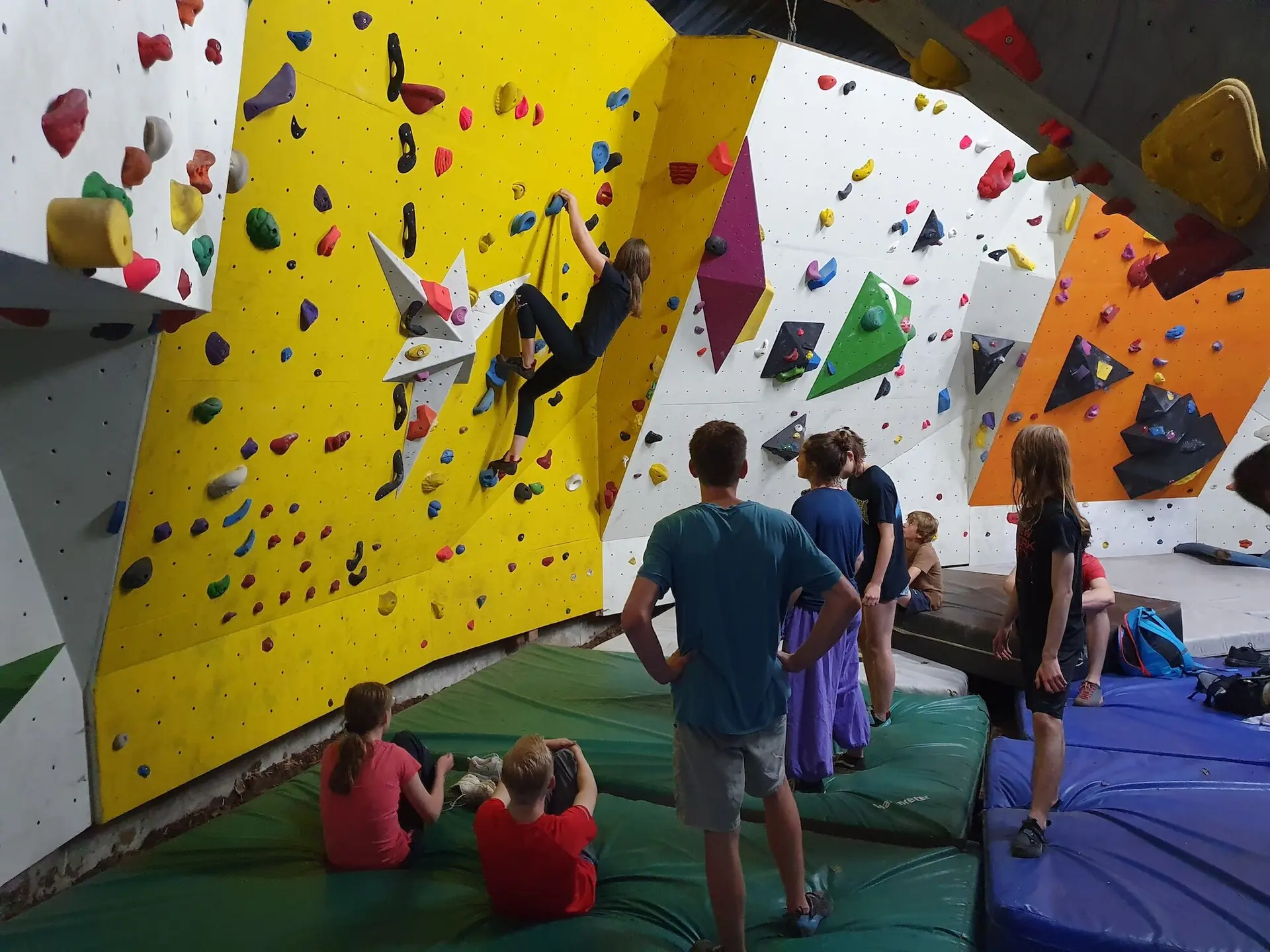 Jugendliche und Betreuer stehen in einer Indoor‑Boulderhalle, einige klettern, andere schauen zu; Gruppenmoment 2023. | © DAV Markt Schwaben