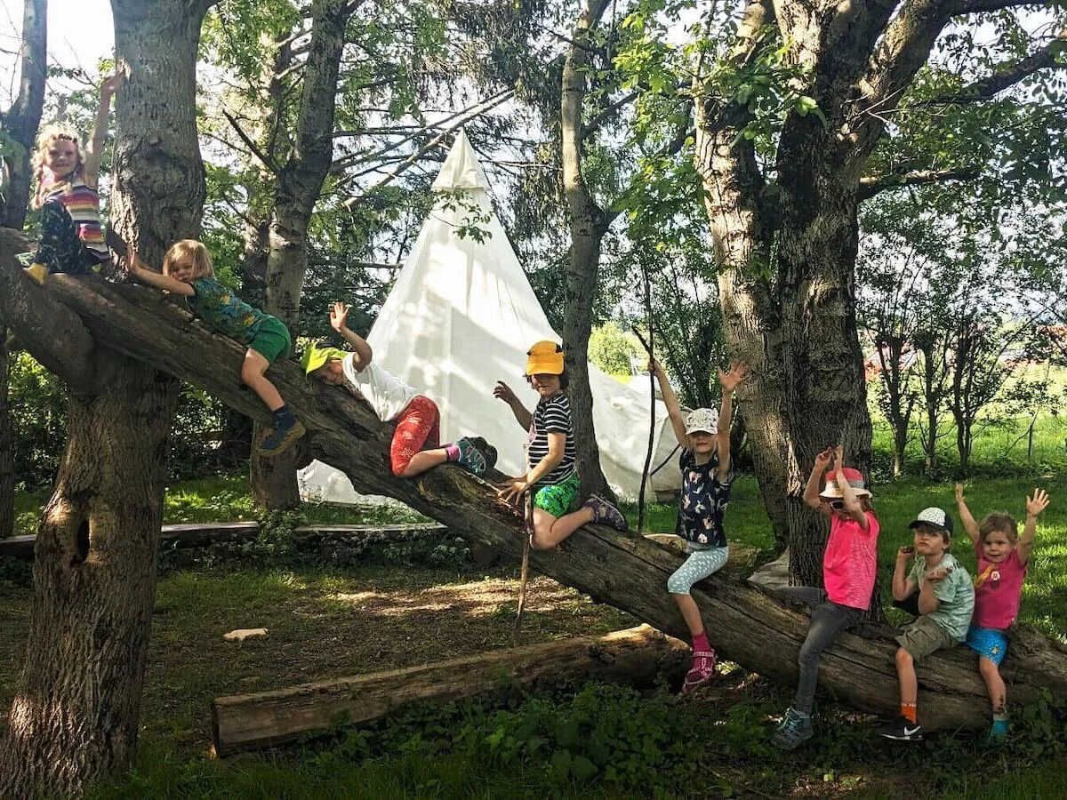 Kinder sitzen fröhlich auf einer Picknickdecke in der Natur und strecken die Arme in die Luft. Umgeben von Rucksäcken und Proviant genießen sie ein gemeinsames Abenteuer – Familiengruppe der DAV-Sektion Markt Schwaben. | © DAV Markt Schwaben