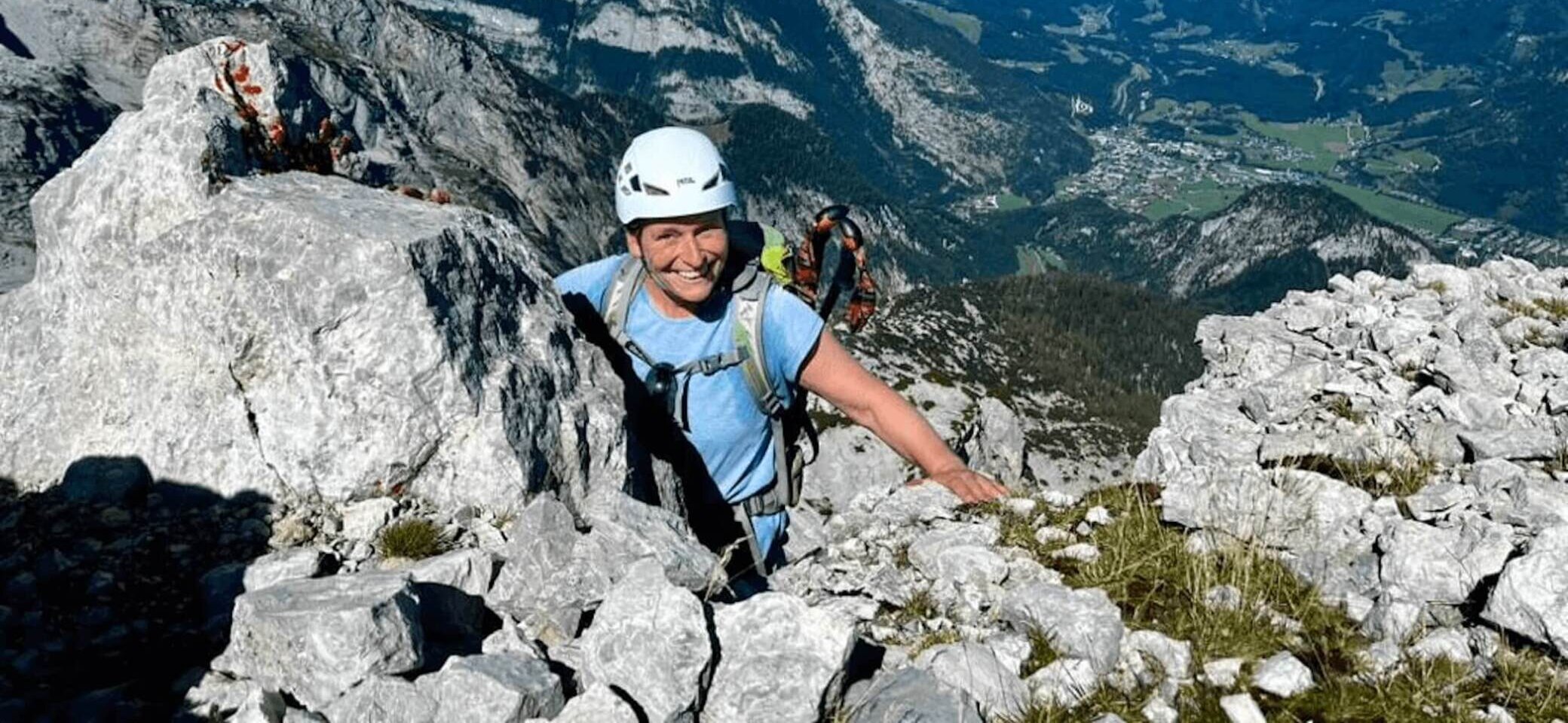 Ein Bergsteiger mit Helm und Rucksack erklimmt eine felsige Wand, während sich im Hintergrund eine weite Landschaft mit Bergen, Tälern und einer kleinen Stadt erstreckt. Das Bild symbolisiert Abenteuer, Herausforderung und die Naturverbundenheit des Alpenvereins. | © DAV Markt Schwaben | Foto Markus Sellmeier