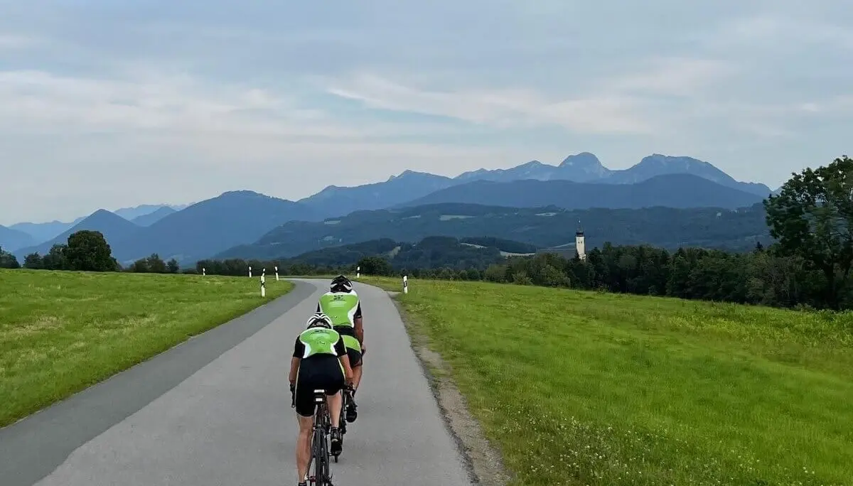 Zwei Radfahrer fahren auf einer Landstraße durch grüne Felder; im Hintergrund Hügel, Berge und ein Kirchturm unter leicht bewölktem Himmel. | © DAV Markt Schwaben