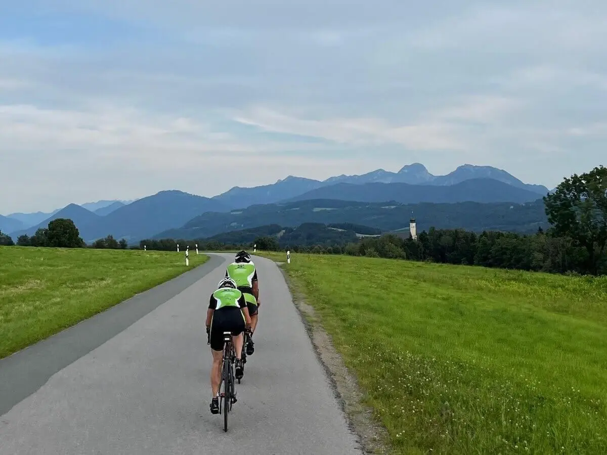Zwei Radfahrer fahren auf einer Landstraße durch grüne Felder; im Hintergrund Hügel, Berge und ein Kirchturm unter leicht bewölktem Himmel. | © DAV Markt Schwaben