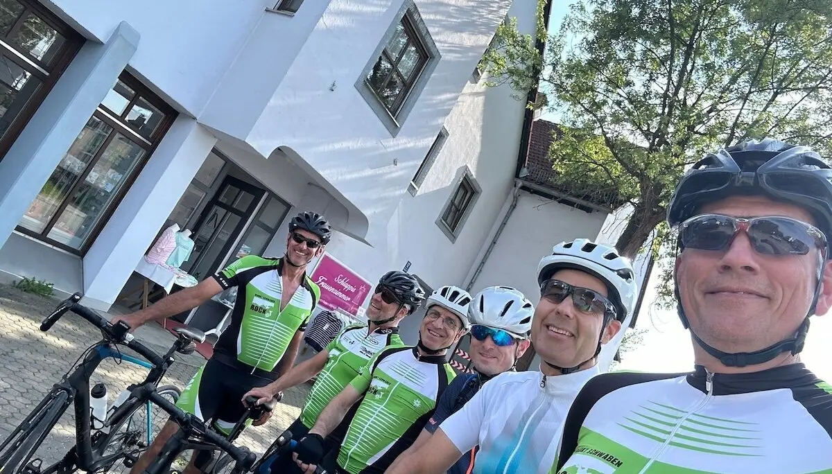 Sechs Radfahrer stehen mit ihren Rennrädern vor einem weißen Gebäude; im Hintergrund Baum, Schilder und weitere Teilnehmende. | © DAV Markt Schwaben