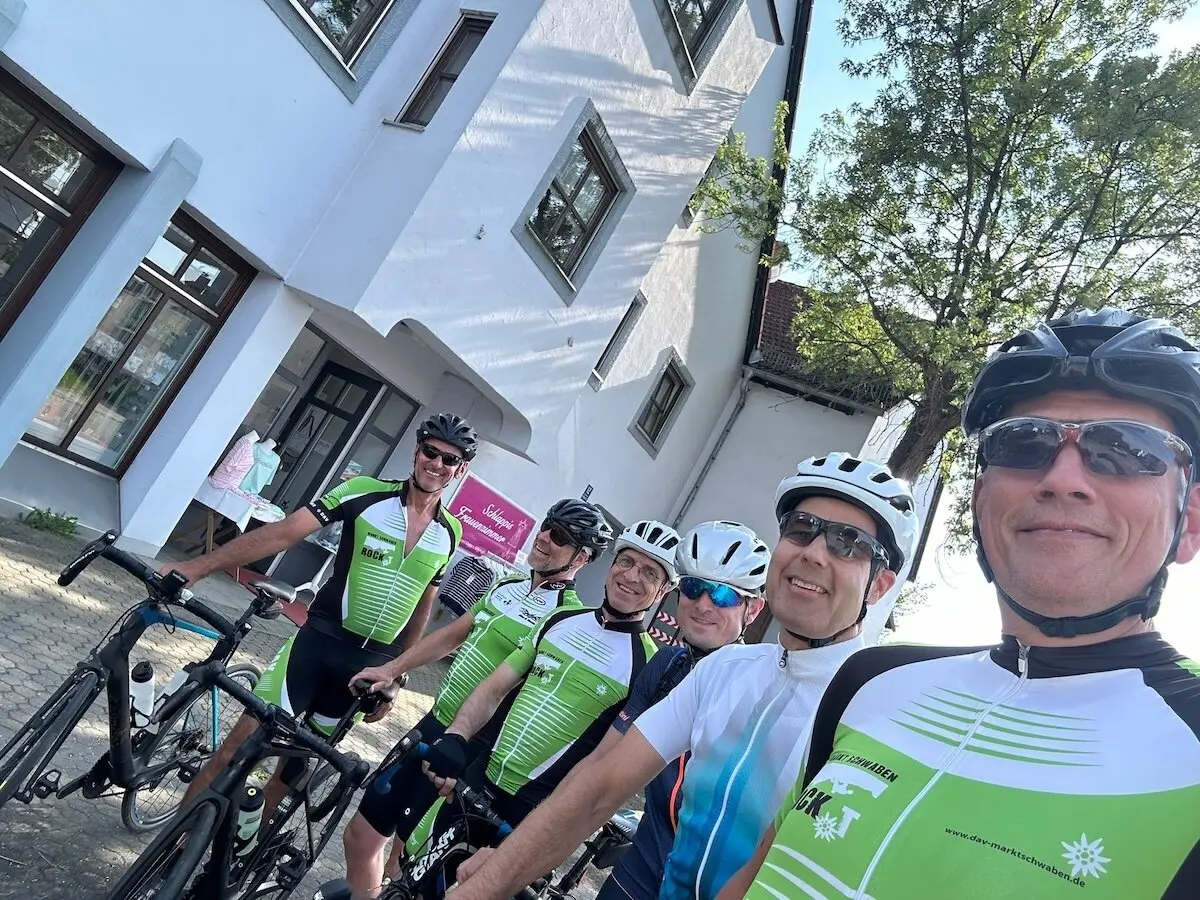 Sechs Radfahrer stehen mit ihren Rennrädern vor einem weißen Gebäude; im Hintergrund Baum, Schilder und weitere Teilnehmende. | © DAV Markt Schwaben