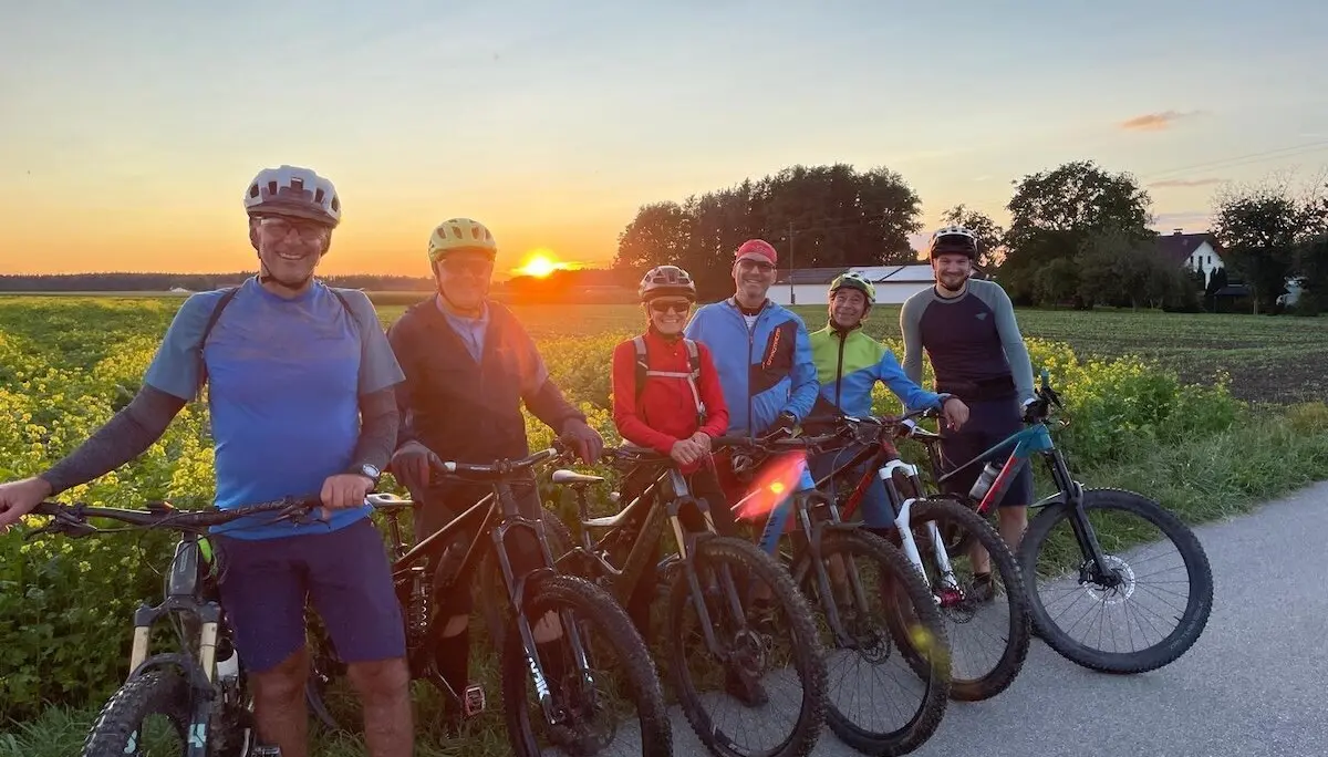 Sechs Radfahrer stehen mit ihren Mountainbikes auf einem Weg neben einem Feld; Sonnenuntergang mit warmem Licht über Landschaft und Häusern. | © DAV Markt Schwaben