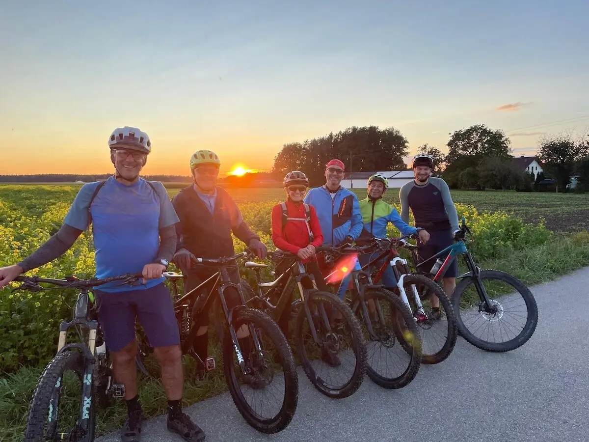 Sechs Radfahrer stehen mit ihren Mountainbikes auf einem Weg neben einem Feld; Sonnenuntergang mit warmem Licht über Landschaft und Häusern. | © DAV Markt Schwaben