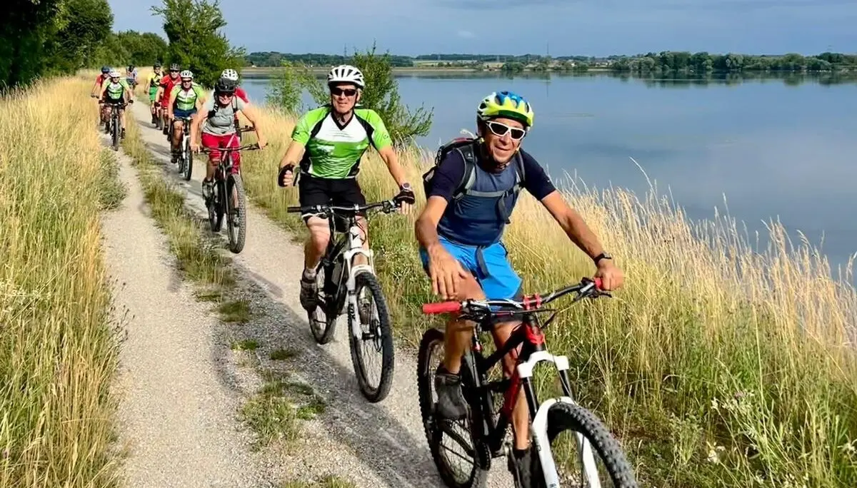 Mehrere Radfahrer fahren auf einem schmalen Kiesweg am See entlang; ruhige Landschaft mit Gras, Bäumen und Wasser unter leicht bewölktem Himmel. | © DAV Markt Schwaben