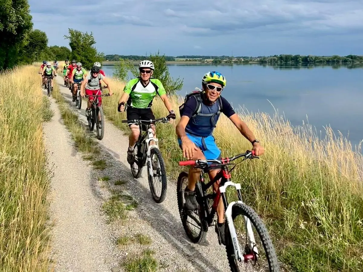 Mehrere Radfahrer fahren auf einem schmalen Kiesweg am See entlang; ruhige Landschaft mit Gras, Bäumen und Wasser unter leicht bewölktem Himmel. | © DAV Markt Schwaben