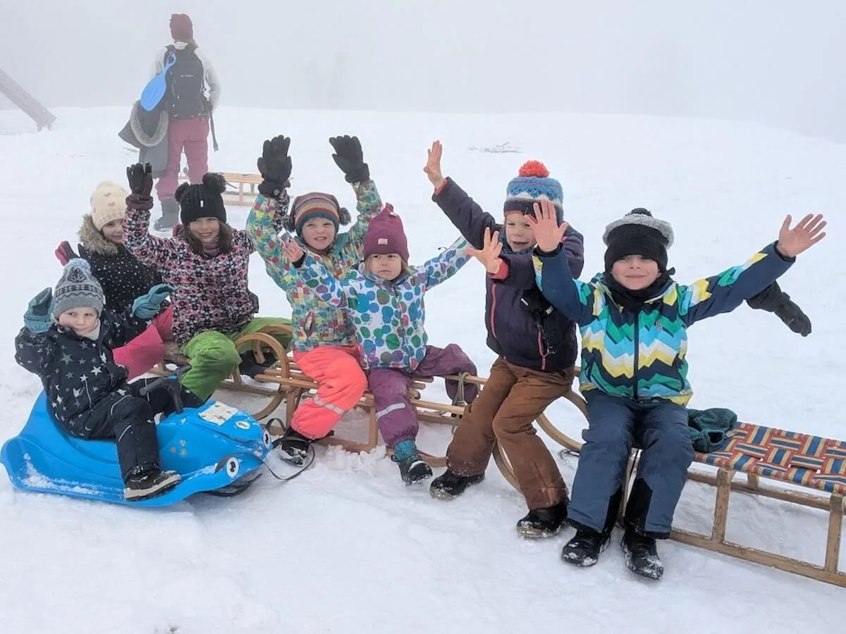Mehrere Kinder sitzen fröhlich auf Schlitten im verschneiten Gelände am Spitzingsee. Sie tragen bunte Winterkleidung und heben jubelnd die Arme. Im Hintergrund steht eine erwachsene Person mit Rucksack und Schlitten. Nebel und Schnee schaffen eine winterliche Stimmung. | © DAV Markt Schwaben