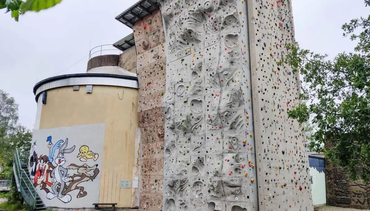 Outdoor‑Kletterwand mit farbigen Griffen an einem Gebäude mit großer Comic‑Bemalung; Holzstämme und Treppe im Vorderbereich. | © DAV Markt Schwaben/Max Mayr