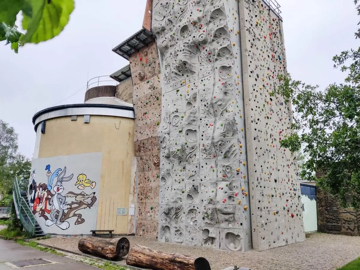 Outdoor‑Kletterwand mit farbigen Griffen an einem Gebäude mit großer Comic‑Bemalung; Holzstämme und Treppe im Vorderbereich. | © DAV Markt Schwaben/Max Mayr