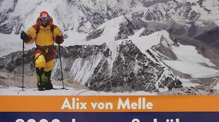 Ein Bergsteiger in gelbem und rotem Anzug steht mit Trekkingstöcken auf einem verschneiten Gipfel. Im Hintergrund schroffe, schneebedeckte Berge. Text: „Alix von Melle – 8000 drunter & drüber – Eine Leidenschaft fürs Leben“. Oben rechts: Logo „GO CLIMB A MOUNTAIN“. | © Alix von Melle