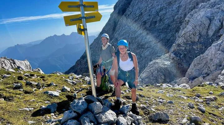 Zwei Wanderer stehen neben einem Wegweiser auf einem Berggipfel. Die gelben Schilder zeigen verschiedene Wanderziele und Gehzeiten in alpiner Umgebung. Diese Szene symbolisiert die Orientierung und Unterstützung, die der DAV Markt Schwaben seinen Mitgliedern bietet – sei es durch Ausrüstung, Verleih, Vereinsartikel, Downloads oder den Newsletter. | © DAV Markt Schwaben