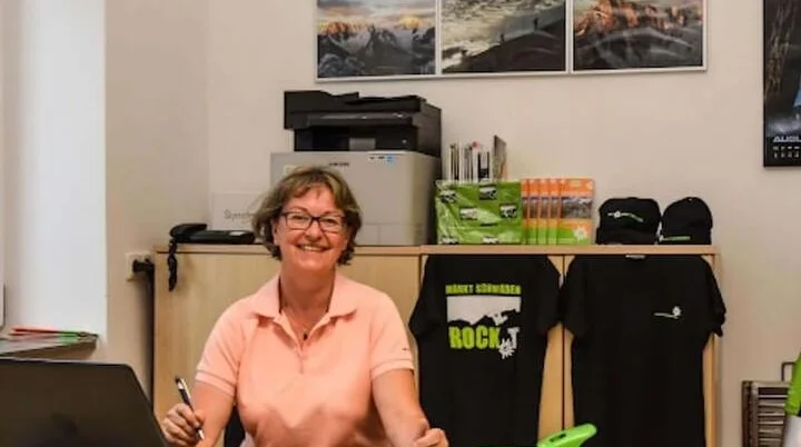 Blick in die Geschäftsstelle des Alpenvereins: Ein Mitarbeiter am Schreibtisch arbeitet am Laptop, während Broschüren, Vereinskleidung und Outdoor-Ausrüstung im Hintergrund präsentiert werden. Jetzt Kontakt aufnehmen und mehr über Mitgliedschaft und Angebote erfahren! | © DAV Markt Schwaben