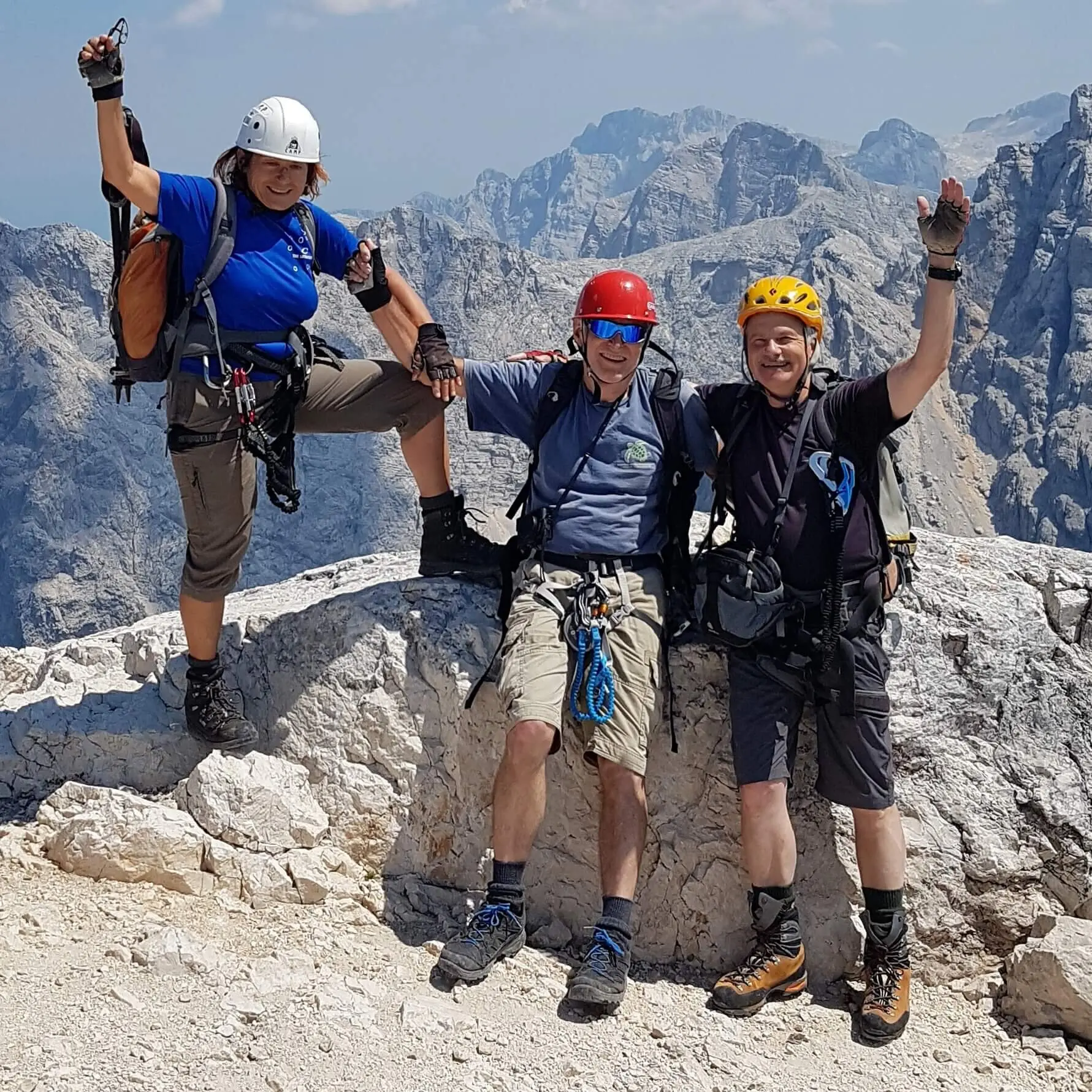 Drei Bergsteiger stehen auf einem felsigen Gipfel, ausgestattet mit Helmen, Klettergurten und Rucksäcken. Hinter ihnen erstrecken sich beeindruckende Bergketten unter einem klaren Himmel – ein Symbol für Abenteuer, Gemeinschaft und das Erreichen gemeinsamer Ziele. | © DAV Markt Schwaben | Foto: Helga Peters