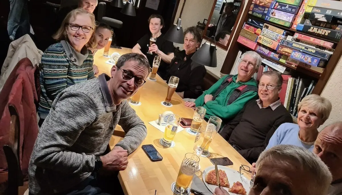 Zwölf Personen sitzen an einem großen Holztisch, trinken Bier und essen Pizza; im Hintergrund steht ein Regal voller Brettspiele. | © DAV Markt Schwaben | Foto Alfred Quiel