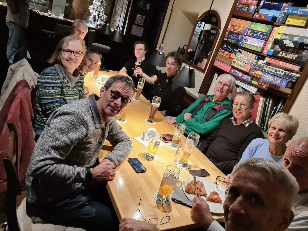 Zwölf Personen sitzen an einem großen Holztisch, trinken Bier und essen Pizza; im Hintergrund steht ein Regal voller Brettspiele. | © DAV Markt Schwaben | Foto Alfred Quiel