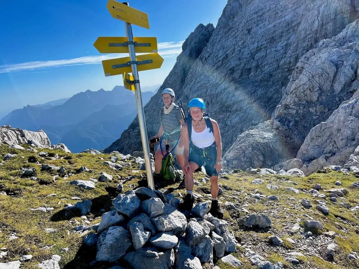 Zwei Wanderer stehen neben einem Wegweiser auf einem Berggipfel. Die gelben Schilder zeigen verschiedene Wanderziele und Gehzeiten in alpiner Umgebung. Diese Szene symbolisiert die Orientierung und Unterstützung, die der DAV Markt Schwaben seinen Mitgliedern bietet – sei es durch Ausrüstung, Verleih, Vereinsartikel, Downloads oder den Newsletter. | © DAV Markt Schwaben