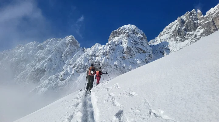 Zwei Bergsteiger steigen mit Schneeschuhen und Stöcken eine verschneite Hangflanke hinauf, umgeben von alpinen Gipfeln und klarem Himmel – Symbol für winterliche DAV-Touren und gemeinsames Naturerleben. | © DAV Markt Schwaben · Fotograf unbekannt