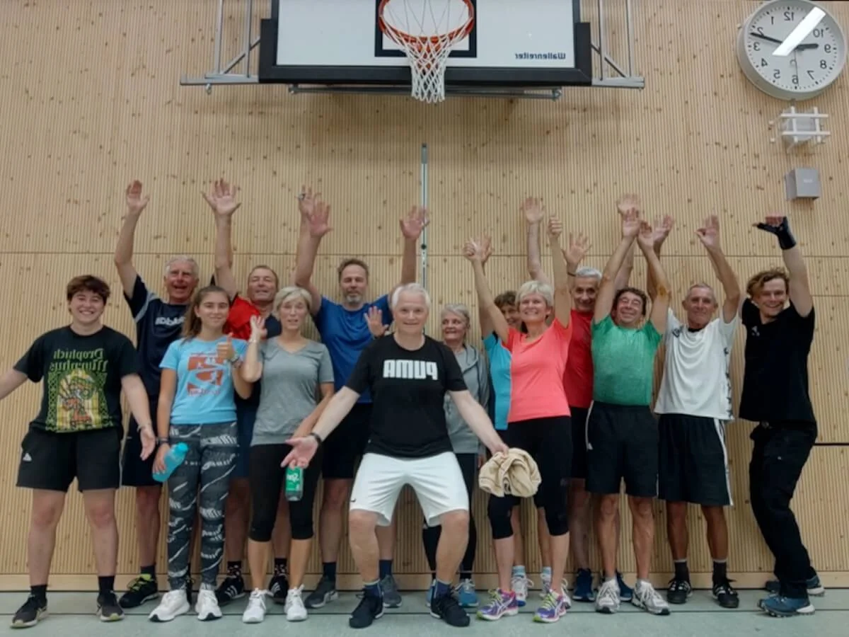 Eine Gruppe von 17 Personen steht in einer Sporthalle unter einem Basketballkorb. Alle tragen Sportkleidung, lächeln in die Kamera und heben die Arme, als gemeinsames Abschlussfoto nach dem Wintertraining. | © DAV Markt Schwaben · Fotograf unbekannt