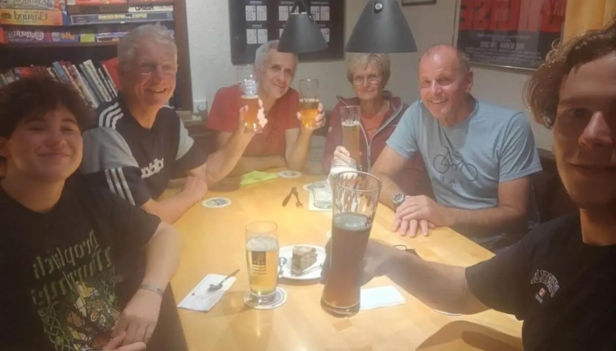 Sechs Personen sitzen an einem Holztisch in einem warm beleuchteten Raum, stoßen mit Biergläsern an und lächeln in die Kamera. Auf dem Tisch stehen Getränke und kleine Snacks, im Hintergrund Regale mit Spielen und Büchern. | © DAV Markt Schwaben · Fotograf unbekannt