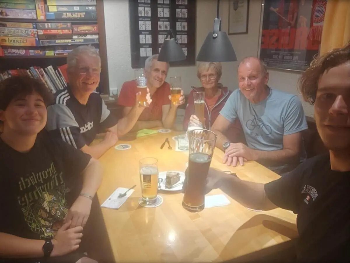 Sechs Personen sitzen an einem Holztisch in einem warm beleuchteten Raum, stoßen mit Biergläsern an und lächeln in die Kamera. Auf dem Tisch stehen Getränke und kleine Snacks, im Hintergrund Regale mit Spielen und Büchern. | © DAV Markt Schwaben · Fotograf unbekannt