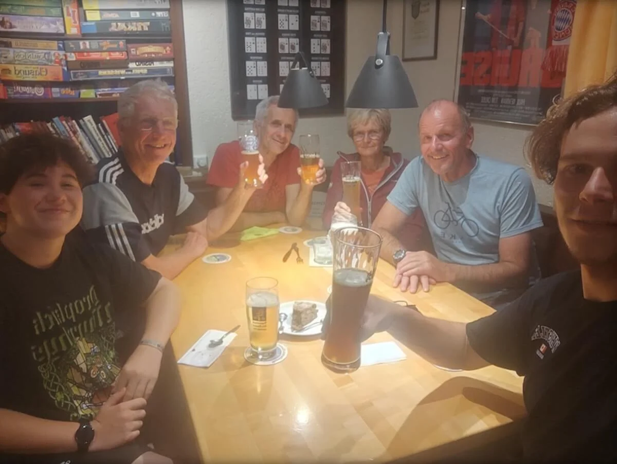 Sechs Personen sitzen an einem Holztisch in einem warm beleuchteten Raum, stoßen mit Biergläsern an und lächeln in die Kamera. Auf dem Tisch stehen Getränke und kleine Snacks, im Hintergrund Regale mit Spielen und Büchern. | © DAV Markt Schwaben · Fotograf unbekannt