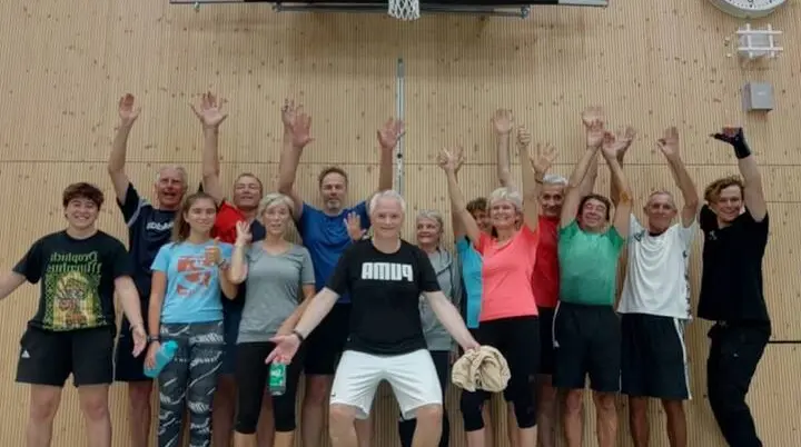 Eine Gruppe von 17 Personen steht in einer Sporthalle unter einem Basketballkorb. Alle tragen Sportkleidung, lächeln in die Kamera und heben die Arme, als gemeinsames Abschlussfoto nach dem Wintertraining. | © DAV Markt Schwaben · Fotograf unbekannt