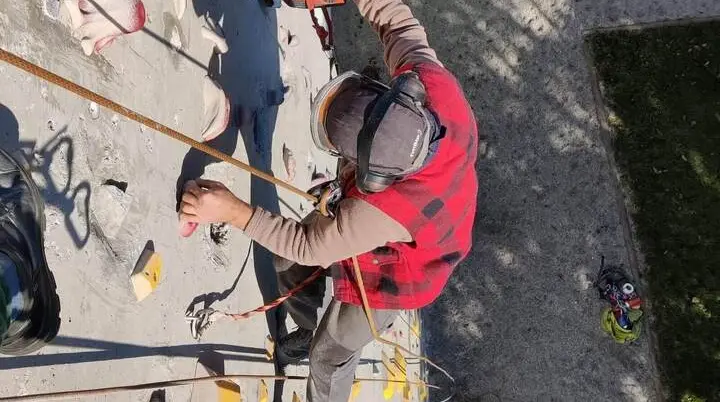 Eine Person sichert sich am Kletterturm und montiert Klettergriffe mit einer Bohrmaschine; sichtbare Schutzausrüstung zeigt den Arbeitsdienst. | © DAV Markt Schwaben / Maximilian Mayr