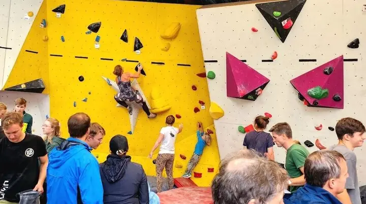 Teilnehmende klettern beim Boulder-Cup an unterschiedlich gestalteten Wandsektionen – gelbe und weiße Flächen mit bunten Griffen, Zuschauer beobachten und feuern an. | © DAV Markt Schwaben