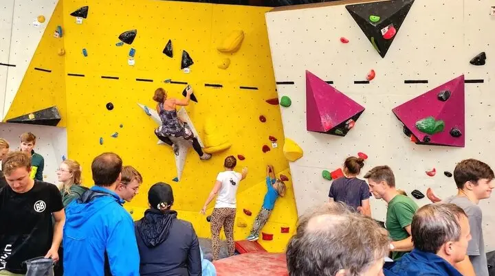 Teilnehmende klettern beim Boulder-Cup an unterschiedlich gestalteten Wandsektionen – gelbe und weiße Flächen mit bunten Griffen, Zuschauer beobachten und feuern an. | © DAV Markt Schwaben