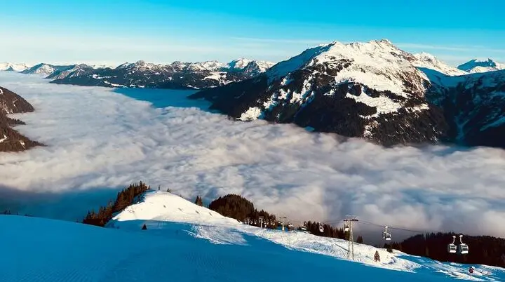 Schneebedeckte Berge mit Skipiste, Sessellift und Wolkendecke im Tal. | © Kerstin Scherrer/Unsplash
